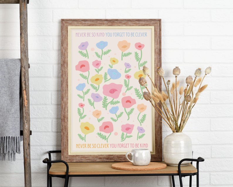 Minimal interior with a light wall, wooden console table, and decorative dried stems, showcasing a framed floral artwork. The design features scattered pastel flowers with soft green stems and text reading “NEVER BE SO KIND YOU FORGET TO BE CLEVER” and “NEVER BE SO CLEVER YOU FORGET TO BE KIND,” creating a gentle, uplifting piece that suits feminine, cozy, or cottagecore-inspired interiors