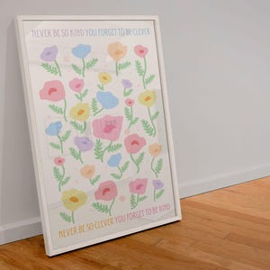 Minimal room with light walls and natural light, featuring a framed floral artwork leaning on the floor. The design shows scattered pastel flowers with green stems and text reading “NEVER BE SO KIND YOU FORGET TO BE CLEVER” and “NEVER BE SO CLEVER YOU FORGET TO BE KIND,” creating a soft, uplifting focal point for feminine, calm, or cozy interior styles