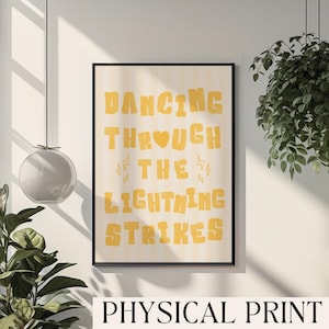 以下が含まれることがあります： 「DANCING THROUGH THE LIGHTNING STRIKES」の黄色い文字がクリーム色の縦縞模様の背景に描かれた、額入りのアートプリント。黒いフレームに入っており、下部には「PHYSICAL PRINT」の文字があります。
