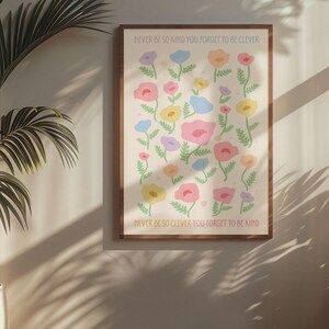 Cozy interior with warm light and minimal decor, displaying a framed floral artwork on the wall. The design features scattered pastel flowers and text reading “NEVER BE SO KIND YOU FORGET TO BE CLEVER” and “NEVER BE SO CLEVER YOU FORGET TO BE KIND,” creating a calming, uplifting piece that works well in feminine, cottagecore, or soft aesthetic spaces