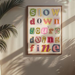 May include: A framed print with the text "slow down you're doing fine" in colorful, cut-out letters. The letters are arranged in a vertical column and are set against a white background.