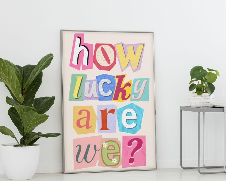 May include: A framed art print with the text "how lucky are we?" in a colorful, cut-out style. The letters are in various colors and fonts, set against a cream background. The print is displayed next to a potted plant.