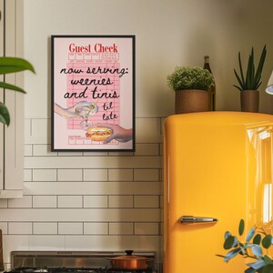 May include: A pink and white framed print with the text "Guest Check" and "now serving: weenies and tinis til late". The print features a cartoon image of a hand holding a hot dog and another hand holding a martini glass.