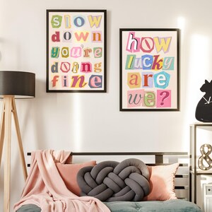 May include: Two framed art prints with colorful text. One reads "SLOW DOWN YOU'RE DOING FINE" and the other says "HOW LUCKY ARE WE?". The prints are in black frames and hang on a white wall above a bed with pillows and a gray knot pillow.