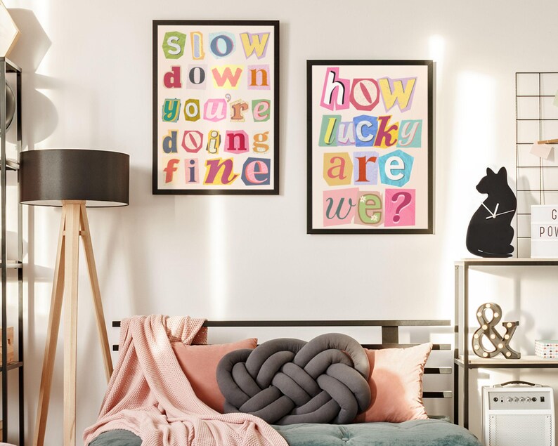 May include: Two framed art prints with colorful text. One reads "SLOW DOWN YOU'RE DOING FINE" and the other says "HOW LUCKY ARE WE?". The prints are in black frames and hang on a white wall above a bed with pillows and a gray knot pillow.