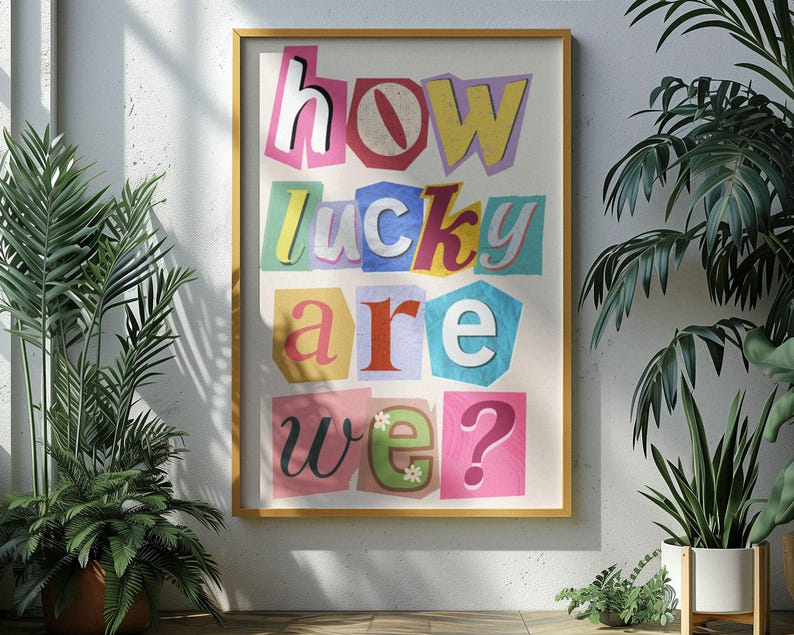 May include: A framed art print with the words "how lucky are we?" in a colorful, cut-out style. The letters are in various colors and fonts, set against a cream-colored background. The frame is a warm, light brown color.