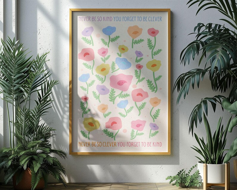 Sunlit room with leafy plants casting shadows across the wall, featuring a framed floral artwork. The design shows soft pastel flowers and green stems with text reading “NEVER BE SO KIND YOU FORGET TO BE CLEVER” and “NEVER BE SO CLEVER YOU FORGET TO BE KIND,” adding a gentle, positive message and soft color palette to modern or nature-inspired interiors