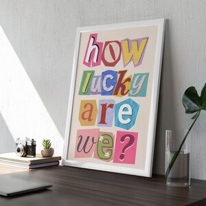May include: Framed art print with the text "how lucky are we?" in a colorful, cut-out style. The artwork features a white frame and is displayed on a desk with a lamp, books, and a small plant. The overall aesthetic is modern and decorative.