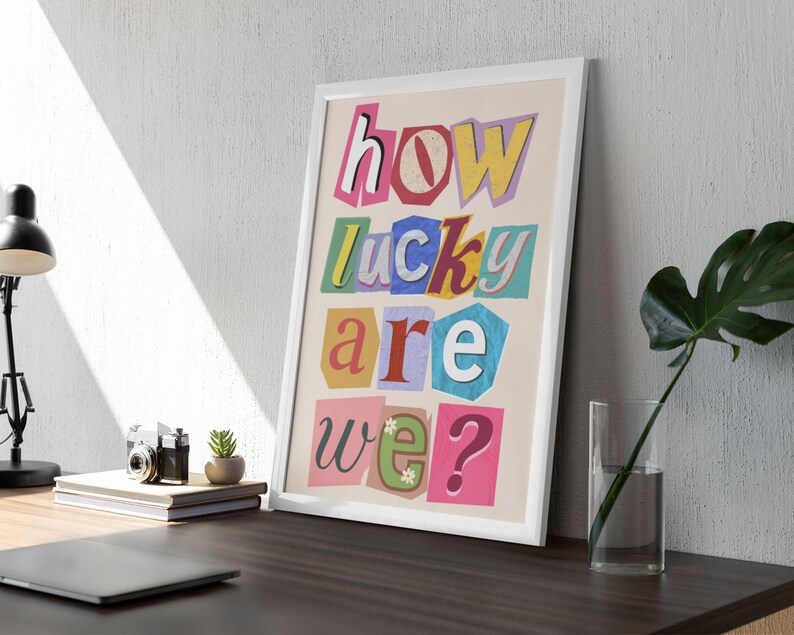 May include: Framed art print with the text "how lucky are we?" in a colorful, cut-out style. The artwork features a white frame and is displayed on a desk with a lamp, books, and a small plant. The overall aesthetic is modern and decorative.