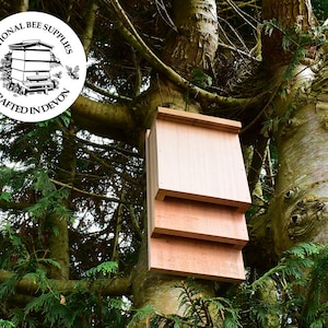 May include: A wooden bat house attached to a tree. The bat house is constructed from light-coloured wood and features a rectangular design with several compartments. The image includes the text "NATIONAL BEE SUPPLIES CRAFTED IN DEVON" within a circular logo.