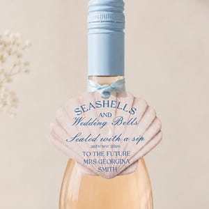 May include: A bottle of Bellini Spritz, an Italian sparkling cocktail, with a light blue cap and a seashell-shaped tag. The tag reads "Seashells and Wedding Bells" and includes a personalized message. The label features a floral design and a glass of the cocktail.