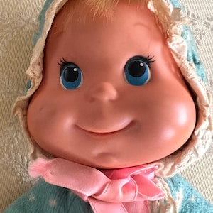 Pode incluir: Uma boneca de plástico com rosto redondo, olhos azuis e cabelo loiro, vestindo uma jaqueta azul-clara com capuz e estampa floral branca. A jaqueta apresenta um acabamento branco com babados ao longo do capuz e uma fita rosa sob o queixo.