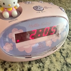 May include: A light pink Hello Kitty digital alarm clock featuring a small Hello Kitty figurine on the left side. The clock has a digital display showing the time 2:05 in red, with a purple floral pattern on the face and a snooze button label.