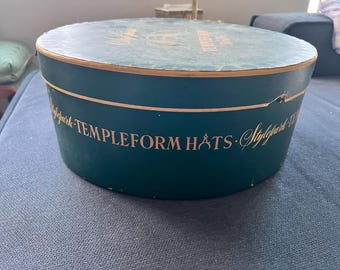 May include: A vintage, round hat box in teal green with gold lettering. The box reads "TEMPLEFORM HATS" and "Stylepark". The lid and rim have a gold trim. The box is sitting on a blue surface.