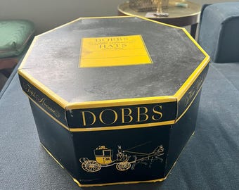 May include: Black octagonal hat box with gold trim and lettering. The box features the text "DOBBS Fifth Avenue HATS" in gold. A gold illustration of a horse-drawn carriage is on the side of the box.