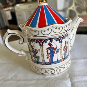 May include: A white ceramic teapot with a circus theme. The lid is striped red, white, and blue, topped with a gold finial. The body features a bandstand scene with musicians and a conductor, accented with gold trim.