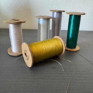May include: A collection of colorful spools of thread. The spools are made of cardboard and hold thread in various colors, including gold, green, silver, and pink. The threads are wound around the spools, and some threads are loose.