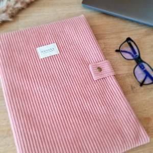 Peut inclure: Une housse d'ordinateur portable en velours côtelé rose avec une étiquette "awenky". La housse a une sangle avec une fermeture à bouton. Des lunettes et un ordinateur portable sont en arrière-plan.