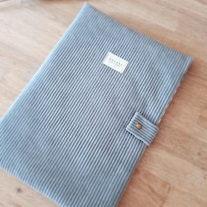 Peut inclure: Une couverture de carnet en velours côtelé bleu clair avec l'inscription "Awenky" sur une petite étiquette. La couverture présente une texture côtelée verticale et un petit rabat avec une fermeture à bouton doré.