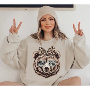May include: Beige sweatshirt featuring a Mama Bear graphic. The bear illustration has a leopard print bow and sunglasses with the text "MAMA BEAR". The model is wearing a beige beanie.