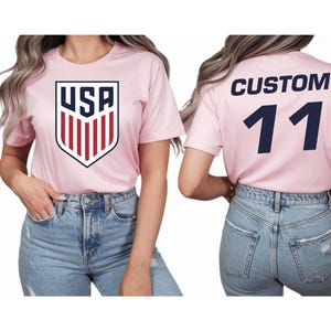 A woman wearing a pink t-shirt with the USA logo and the number 11 on the back, paired with blue jeans.
