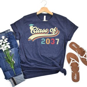 May include: A navy blue t-shirt with the text "Class of 2037" in a retro design. The shirt is paired with blue jeans, white flowers, and brown sandals. The t-shirt is hanging on a wooden hanger.