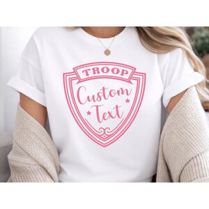 A person wearing a white T-shirt with a pink shield-shaped logo that says "TROOP Custom Text".