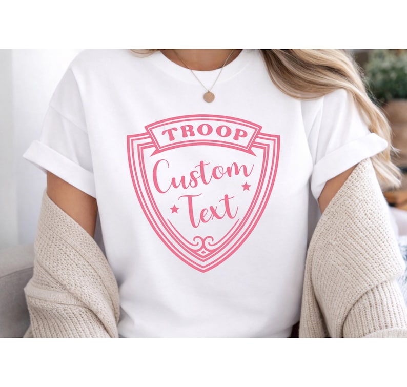 A person wearing a white T-shirt with a pink shield-shaped logo that says "TROOP Custom Text".