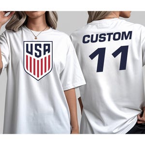 A white t-shirt with a large shield-shaped logo and the text "USA" printed on it, along with the number "11" printed below.