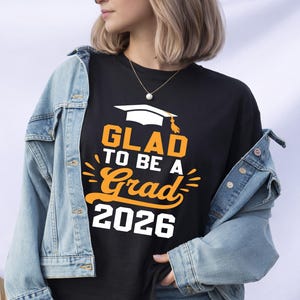 A person wearing a black t-shirt with the text "GLAD TO BE A GRAD 2026" printed on it, along with a graduation cap graphic.