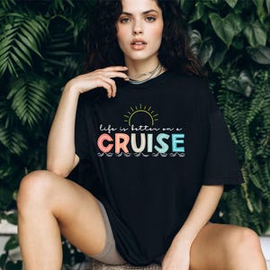 May include: Black t-shirt with the text "life is better on a CRUISE" in colorful letters, a yellow sun graphic, and wavy lines. The shirt is worn by a person sitting in front of a green leafy background.