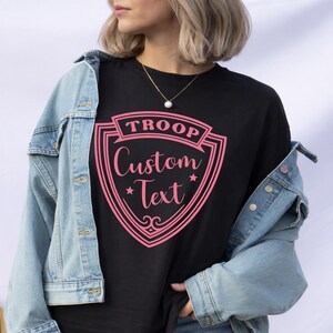 A woman wearing a black t-shirt with a pink shield-shaped graphic and a denim jacket, standing against a white background.