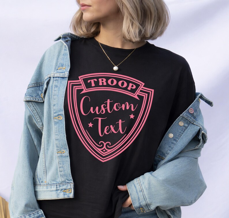 A woman wearing a black t-shirt with a pink shield-shaped graphic and a denim jacket, standing against a white background.