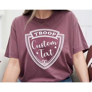 A person wearing a maroon t-shirt with a shield-shaped design that says "TROP Custom Text" printed on it.