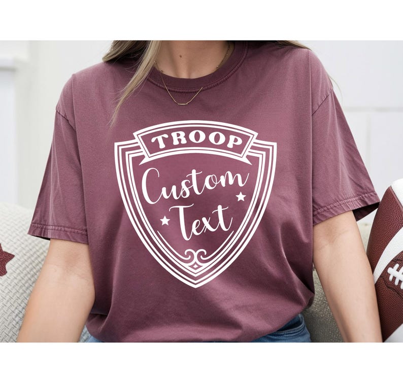 A person wearing a maroon t-shirt with a shield-shaped design that says "TROP Custom Text" printed on it.