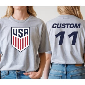 A woman wearing a grey t-shirt with a large shield-shaped logo and the text "USA" on it, as well as the number "11" on the back.