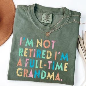 May include: Olive green Comfort Colors t-shirt with the text "I'M NOT RETIRED I'M A FULL-TIME GRANDMA" in colorful letters. A gold necklace with a pendant is draped on the shirt.
