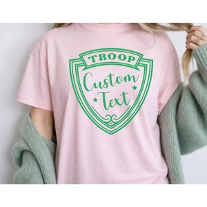 A person wearing a pink t-shirt with a green shield-shaped logo that says "Troop Custom Text".