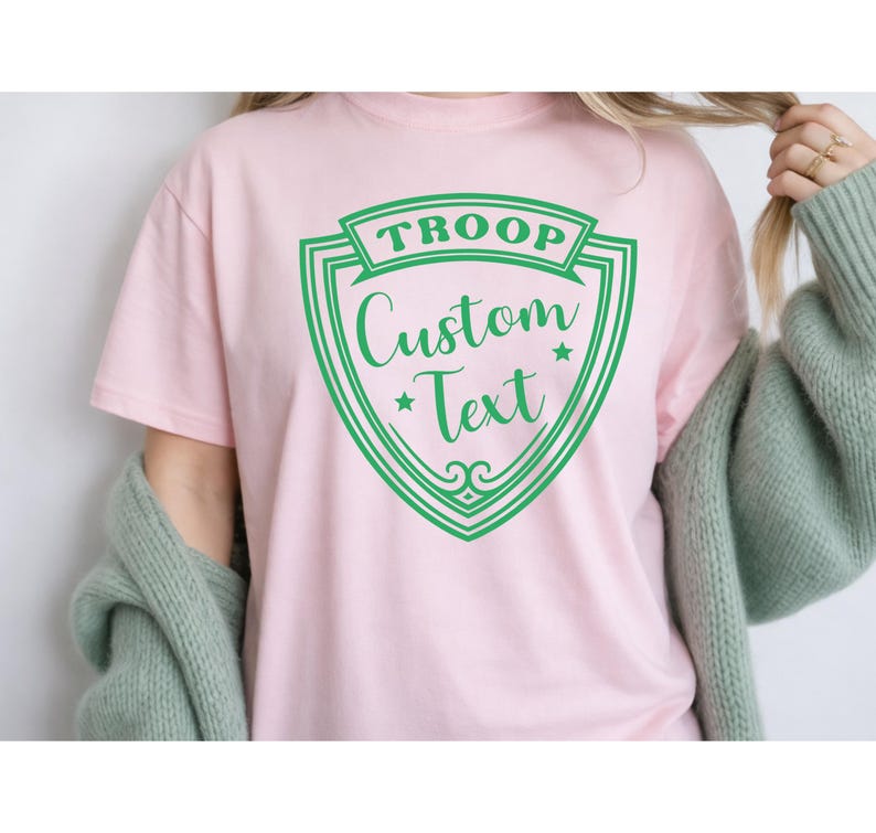 A person wearing a pink t-shirt with a green shield-shaped logo that says "Troop Custom Text".