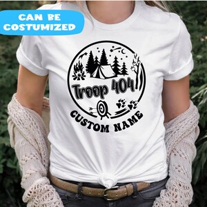 May include: White t-shirt with a black circular graphic featuring a tent, trees, campfire, stars, and the text "Troop 404 Custom Name." The shirt also has the text "Can Be Costumized" in a blue banner.