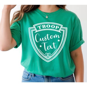 A person wearing a green T-shirt with a shield-shaped design and text printed on it.