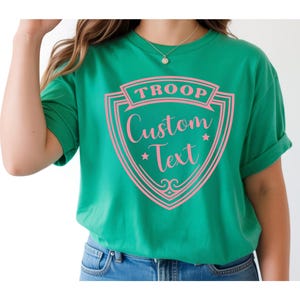 May include: A green t-shirt with a pink shield design. The shield has the word "TROOP" at the top and "Custom Text" in a script font. Two small stars and decorative flourishes are also included in the design.