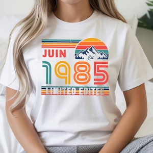 May include: A white short-sleeve t-shirt featuring a graphic with a retro sunset, mountain silhouette, and the text JUIN 1985 LIMITED EDITEE. The design uses horizontal stripes in orange, yellow, teal, and blue, with the year 1985 in large font.