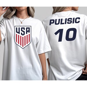 A white t-shirt with a red and blue shield-shaped logo and the name 'PULISIC' printed on the back, along with the number '10'.