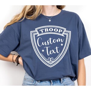 A person wearing a navy blue t-shirt with a white shield design and text printed on it.