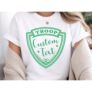 A person wearing a white T-shirt with a green shield-shaped logo that says "TROOP Custom Text".