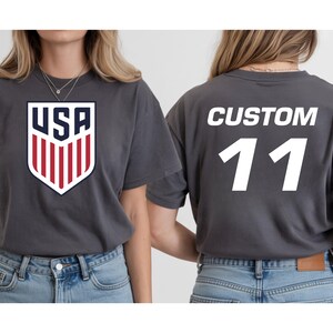 A woman wearing a grey t-shirt with a large shield-shaped logo and the text "USA" on it, and a custom number "11" on the back.