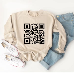 May include: A beige sweatshirt with a large black QR code design. The sweatshirt is paired with light blue jeans and white canvas sneakers. The sneakers have red trim and the word "CONVERSE" printed on the side.