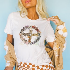 以下が含まれることがあります： 花柄の平和マークがプリントされた白いTシャツ。平和マークは、ピンク、イエロー、オレンジ、グリーンの色合いの様々な花と葉で構成されています。