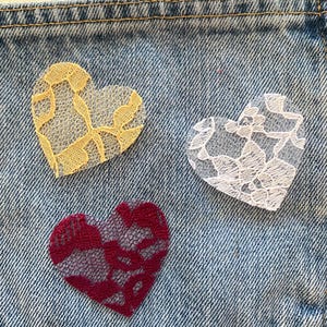 May include: Three heart-shaped lace appliqués attached to a blue denim fabric surface. The hearts are colored yellow, white, and deep red, each featuring a distinct floral lace pattern with visible mesh detailing and stitched edges.